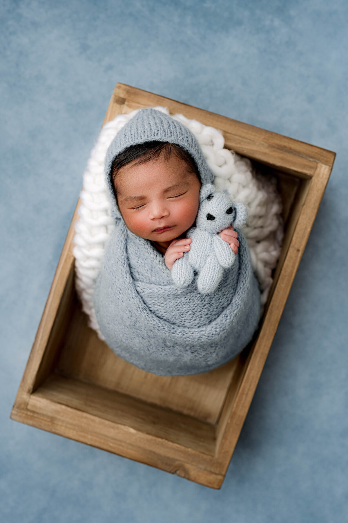 Newborn baby wrapped in a blue knit swaddle and bonnet, sleeping in a wooden crate on a fluffy white blanket, holding a tiny matching teddy bear.
