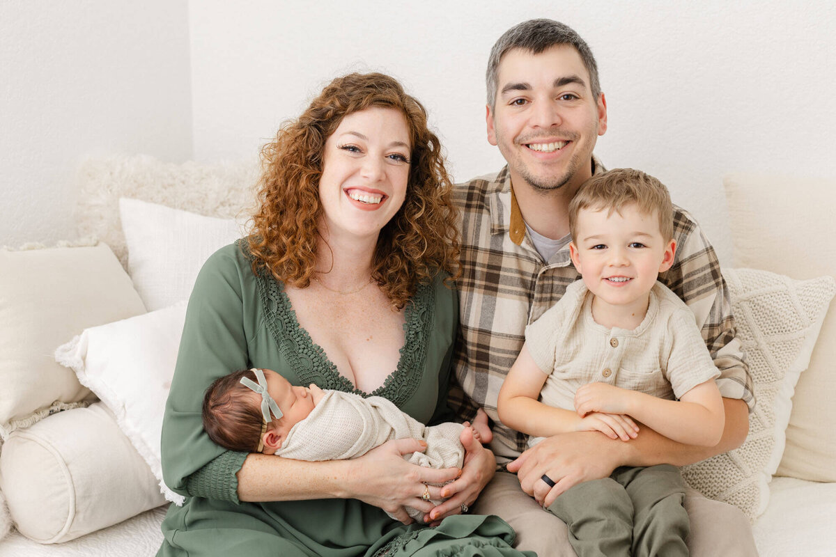 Mom holding newborn baby while dad holds toddler brother. Mom, Dad and Toddler are all smiling at the camera while baby is fast asleep during newborn photos in PDX newborn photography studio.