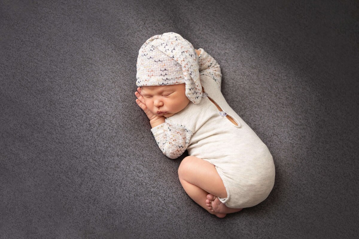 baby wearing a white outfit and hat curled up on grey blanket
