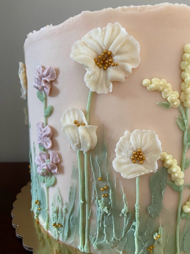 A close up shot of the side of a cake shows a pink base, with buttercream piped florals like a field of wildflowers in white, purple, and yellow with gold accents and green buttercream swatches. 