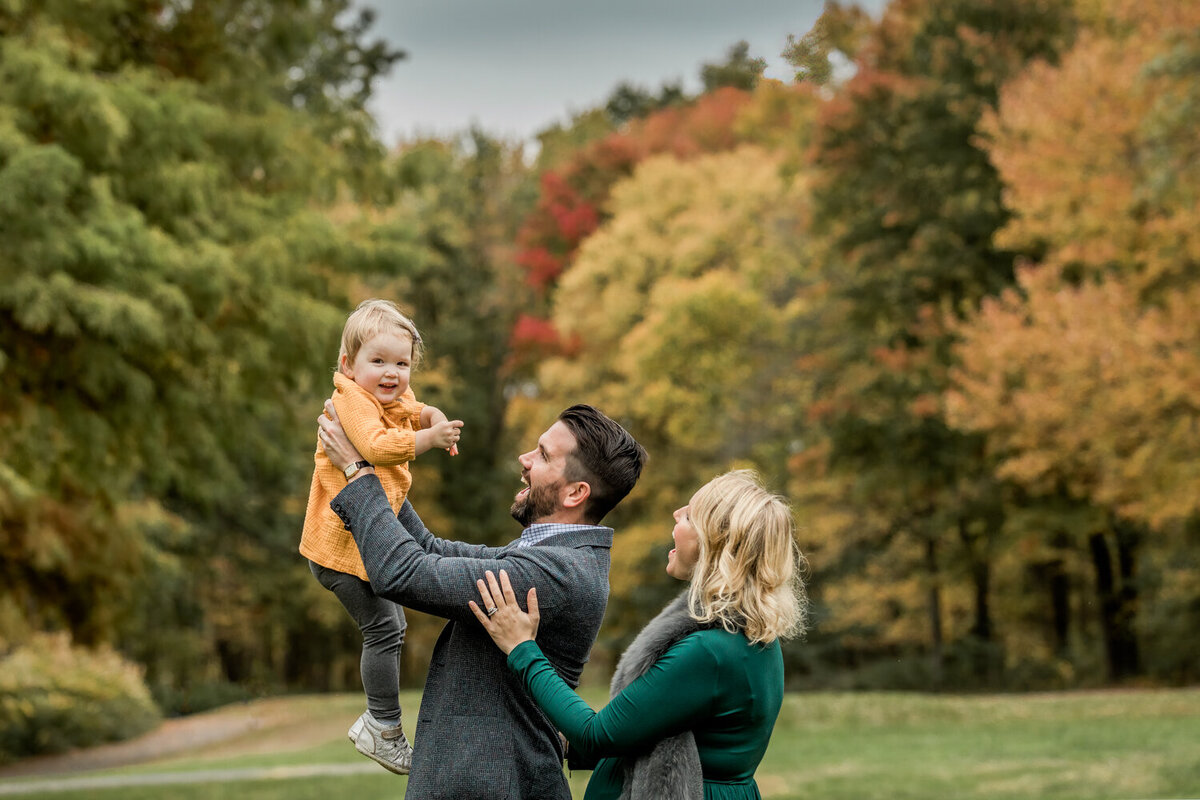 NJ Fall family photo of dad holding toddler girl in the air and mother watching in Oak Ridge Park, Clark, NJ