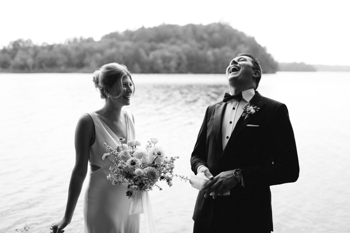 Black and white photo of a bride holding flowers and a groom laughing by a lake, with trees in the background in Muskoka, captured by Jennifer van Son, a wedding photographer in Toronto.