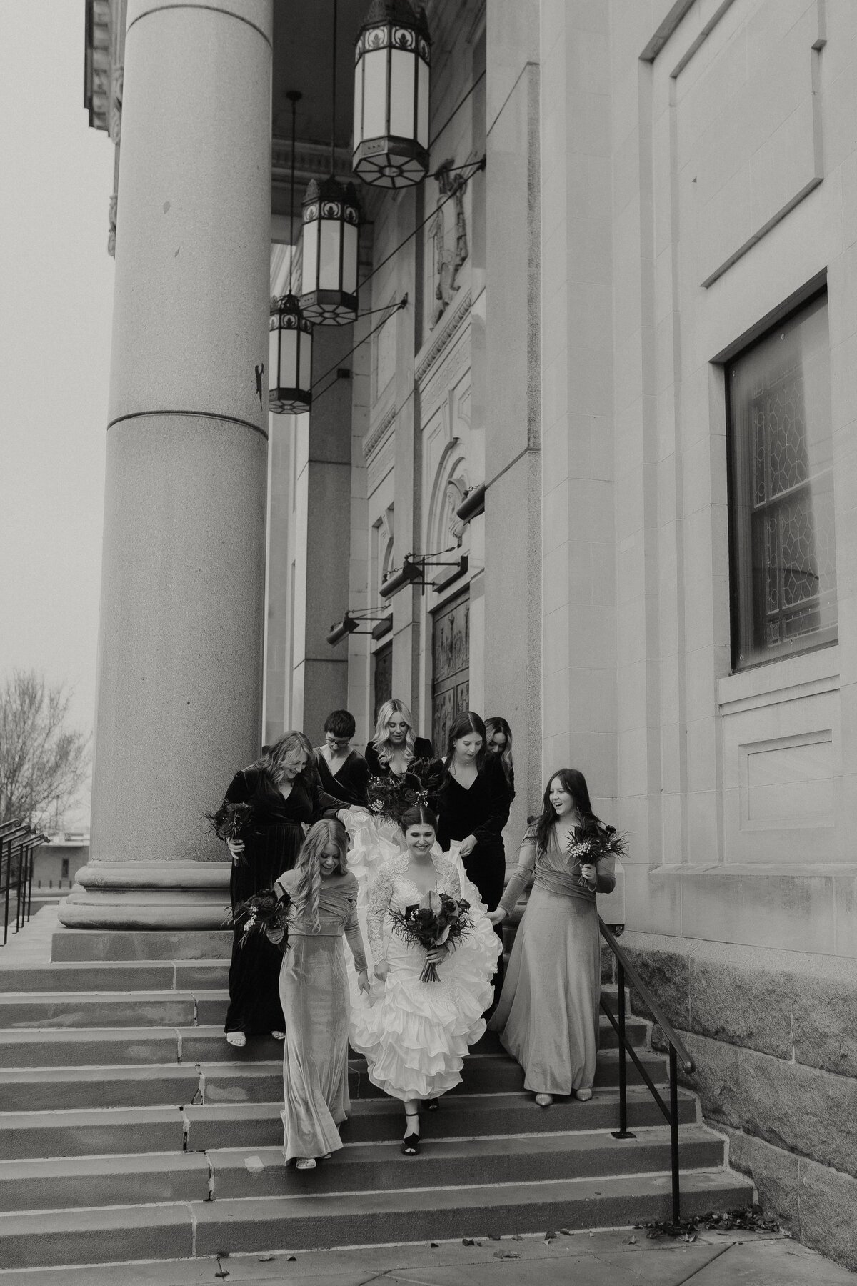 bridal party holding dress while bride walks down stairs