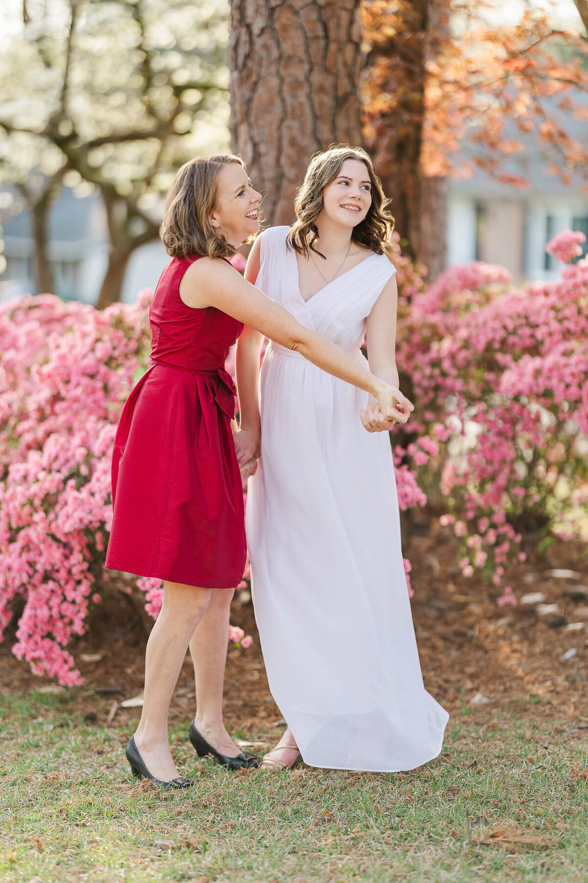 Two women stand outdoors in front of blooming pink azaleas and a tree, captured perfectly by a Fayetteville NC family photographer. One is in a red dress, laughing, while the other wears a flowing white dress, both smiling as they enjoy the spring day.