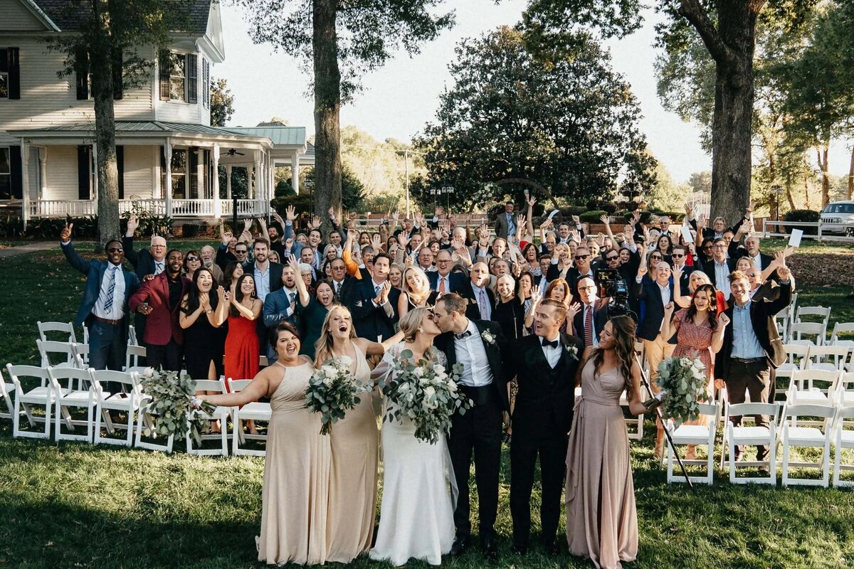 A large wedding party and guests cheering and raising their hands in front of a historic house and white chairs