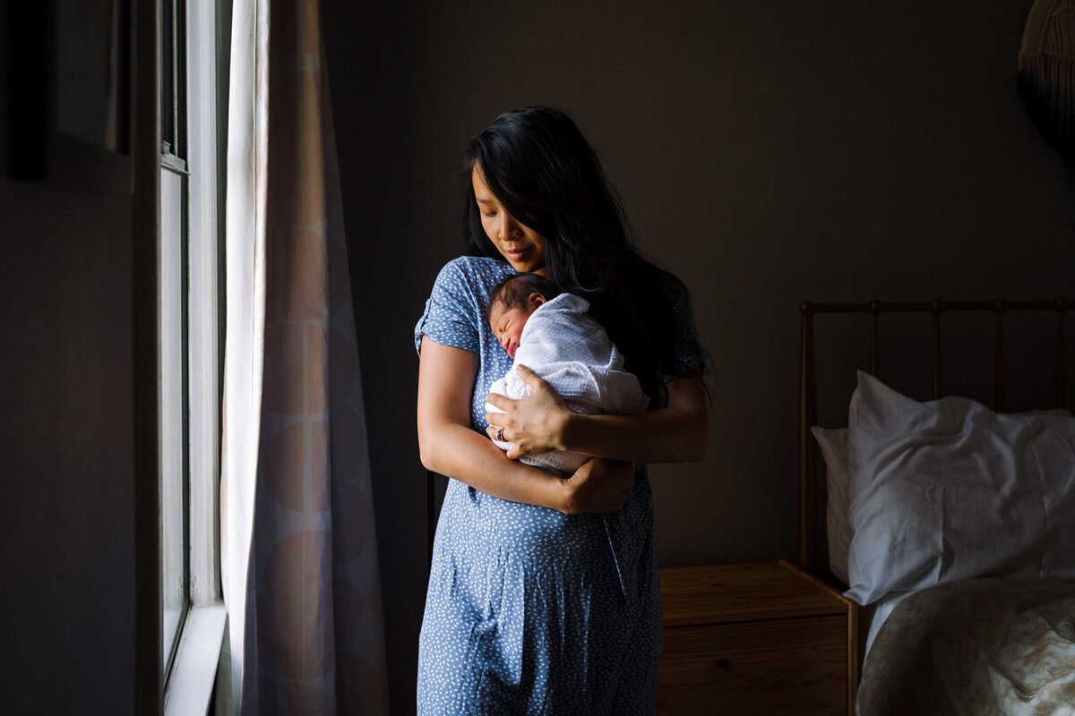 mom in blue dress standing by window holding newborn wrapped in white swaddle