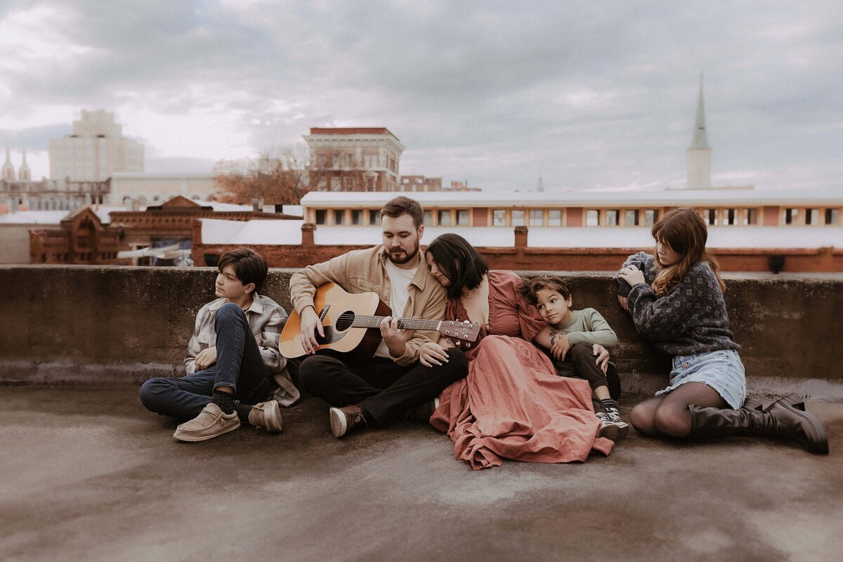 A group of five people sits on a rooftop. A person in the center plays an acoustic guitar. Two children sit nearby, while another person sits on the right, facing away. The background shows city buildings.
