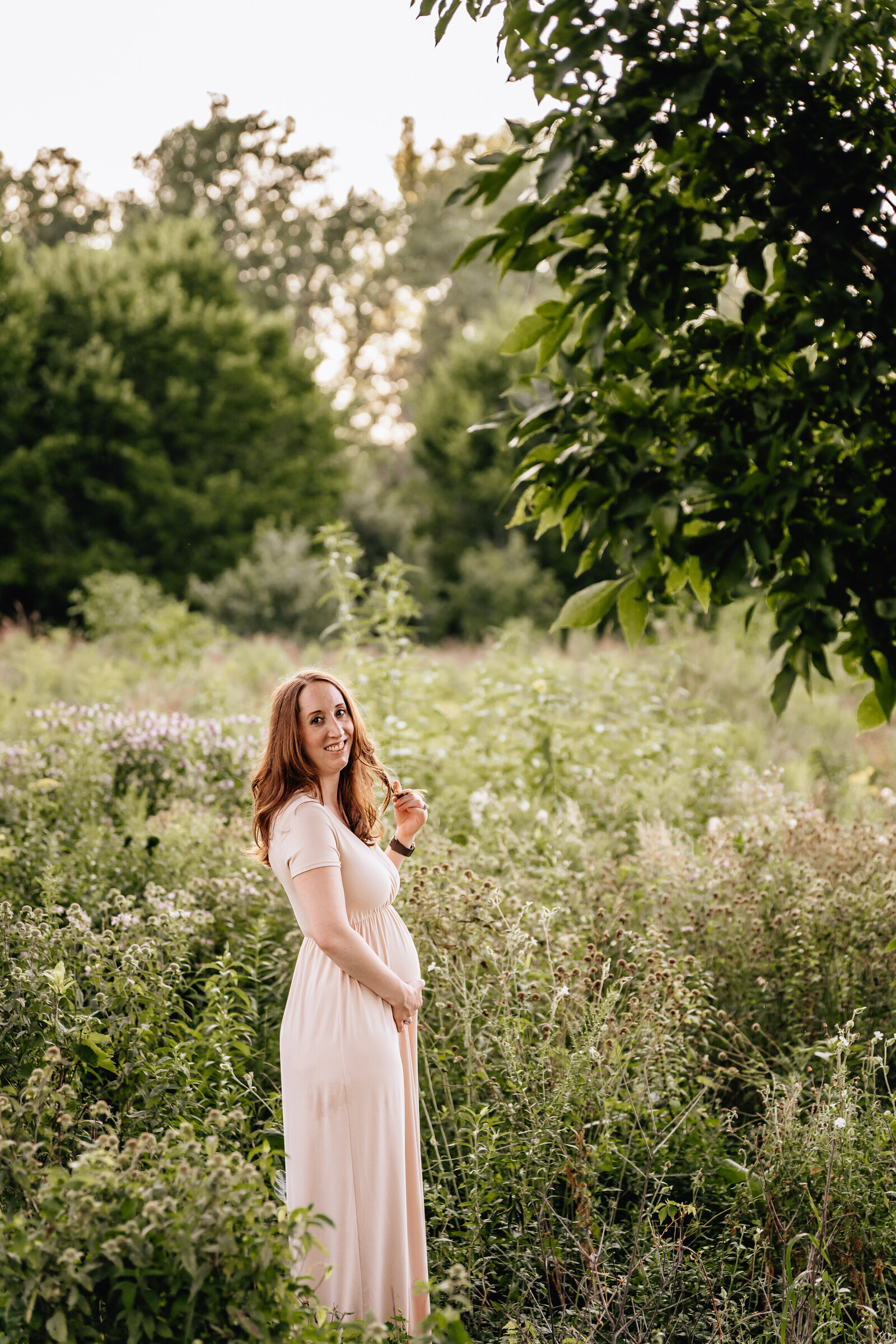 A pregnant woman in a light peach dress stands in a field of wildflowers, smiling gently as she touches her belly. The sun sets behind her, casting a soft golden light over the lush green surroundings.