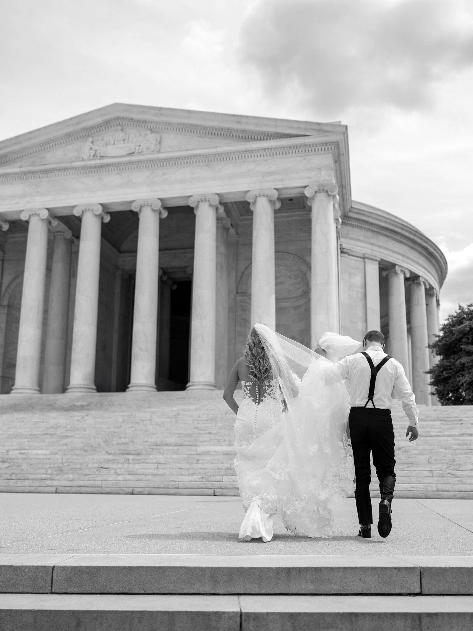 A newlywed couple walks hand in hand towards a monument. The bride wears a flowing white gown and the groom dons a suit with suspenders. The scene is captured in black and white.