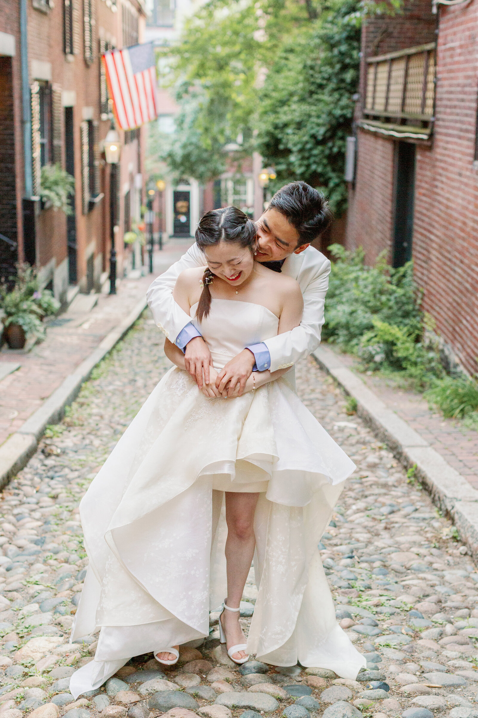 A groom hugging their bride from behind while standing on a small cobblestone road.