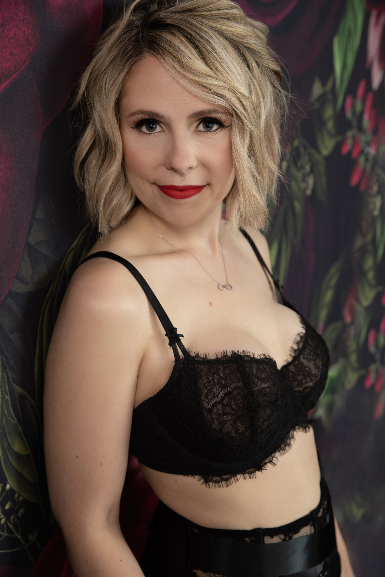 A woman with short, wavy blonde hair is dressed in a black lace bra and black bottoms. She stands against a dark, floral background and looks directly at the camera with a confident expression, wearing red lipstick and a delicate necklace.