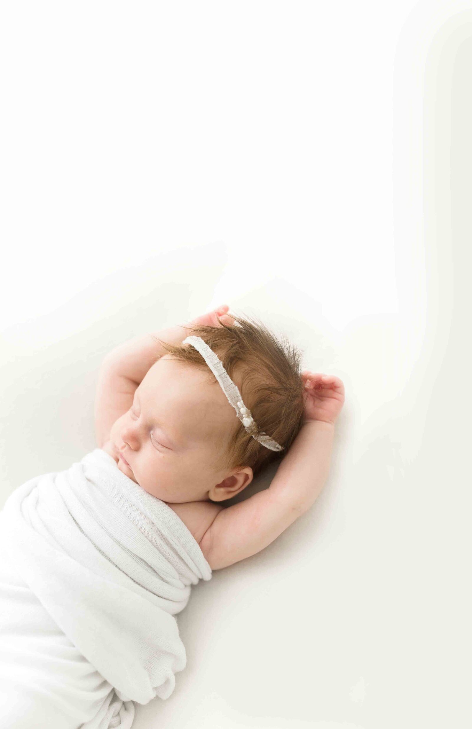 Schlafendes neugeborenes Mädchen mit ausgestreckten Armen in weißer Decke und mit weißem Haarband.