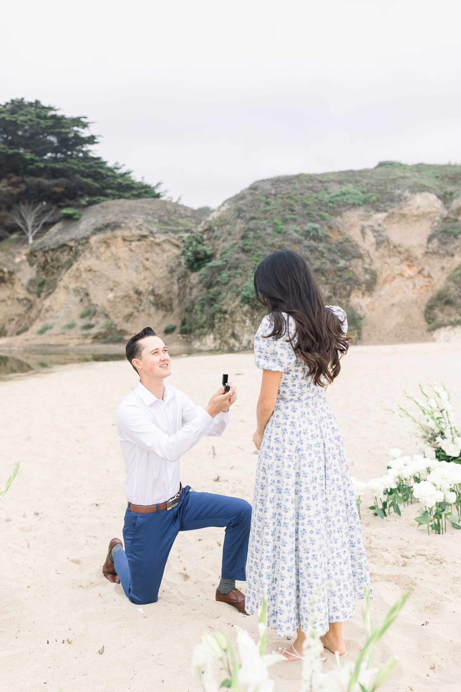 man on one knee at Half Moon Bay with a little box that holds a ring for proposal pictures captured by wedding photographer bay area with woman in a white and blue dress with white flowers around them