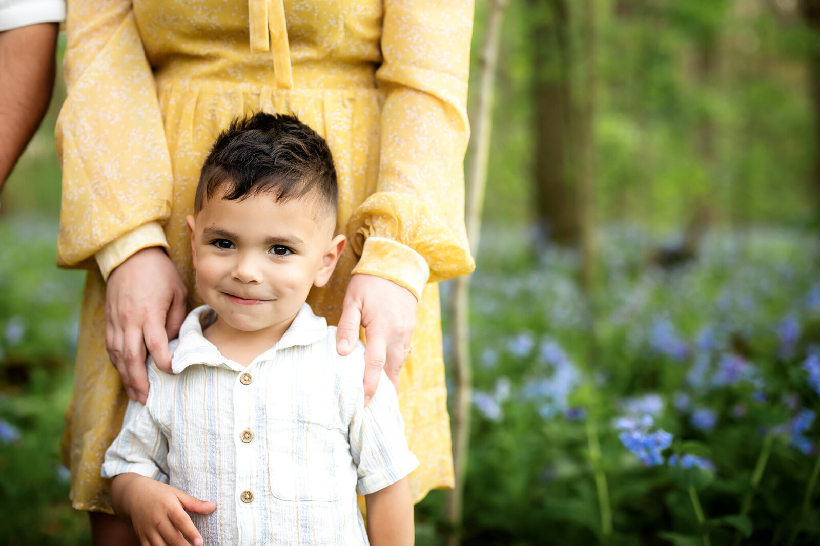 A young boy stands in front of an adult wearing a yellow dress, surrounded by green foliage and purple-blue wildflowers in a sunlit forest. Capturing joyful moments, this Bloomington family photographer specializes in outdoor portraits.