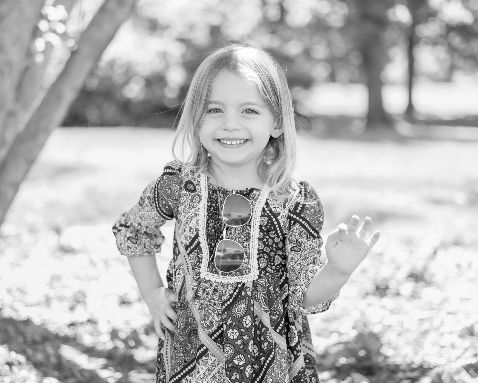 black and white photo of a young girl wearing a print dress, sunglasses resting on her dress, with a hand on her hip, smiling at the camera