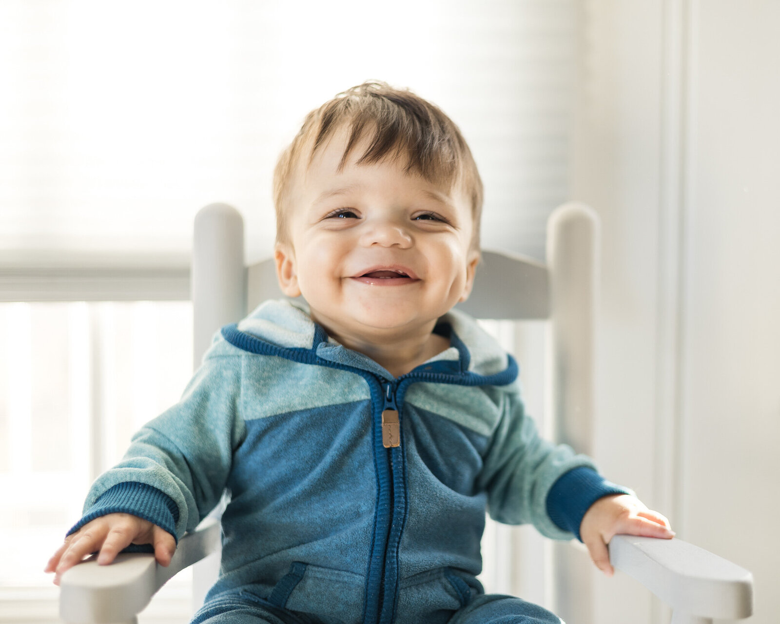 A 1 year old boy with dark brown hair sitting in a light gray rocking chair, with his hands together, looking off camera smiling, wear a blue zip up outfit