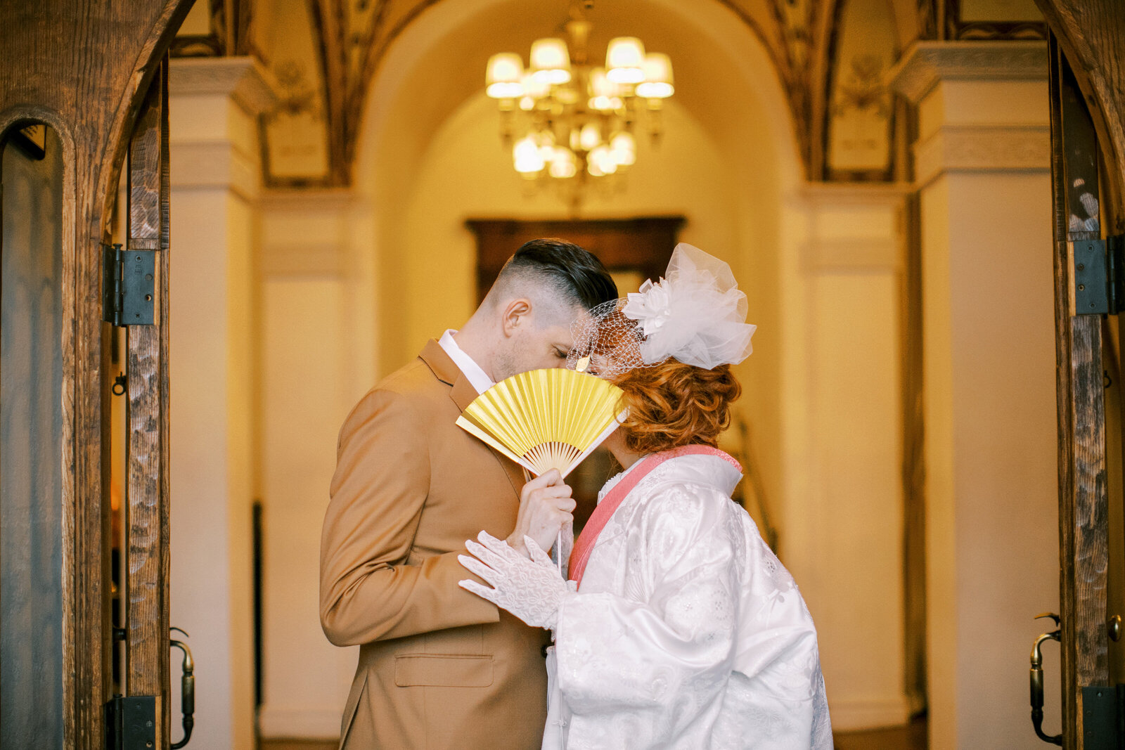 Bride and groom sharing a romantic moment behind a golden fan, bride wearing white shiromuku kimono with pink accents at Carondelet House, Los Angeles California, perfect for wedding kimono rental | 金扇子越しに見つめ合う白無垢ピンク掛下の花嫁と新郎 カロンデレットハウス ロサンゼルス カリフォルニアで着物レンタル