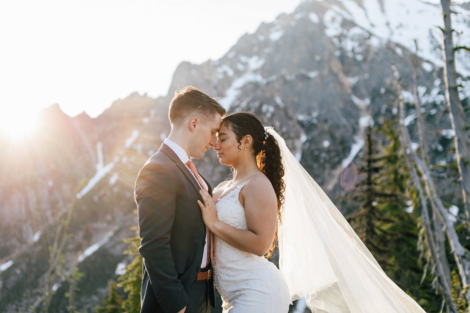 A bride, with long straight brown hair, holds a slice of cake on one hand while kissing her blond haired, grey jacket groom with the forest and Sparks Lake surrounding them at their Bend Oregon elopement. | Erica Swantek Photography