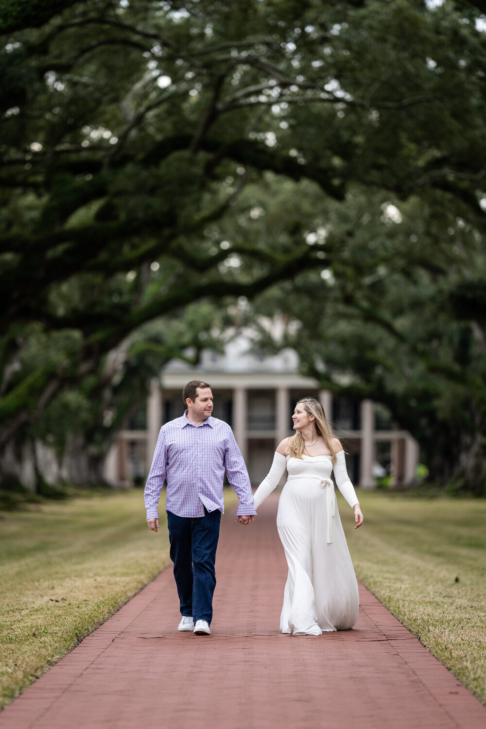 Maternity portrait of couple at Oak Alley Plantation, walking down brick pathway with trees and house in the background