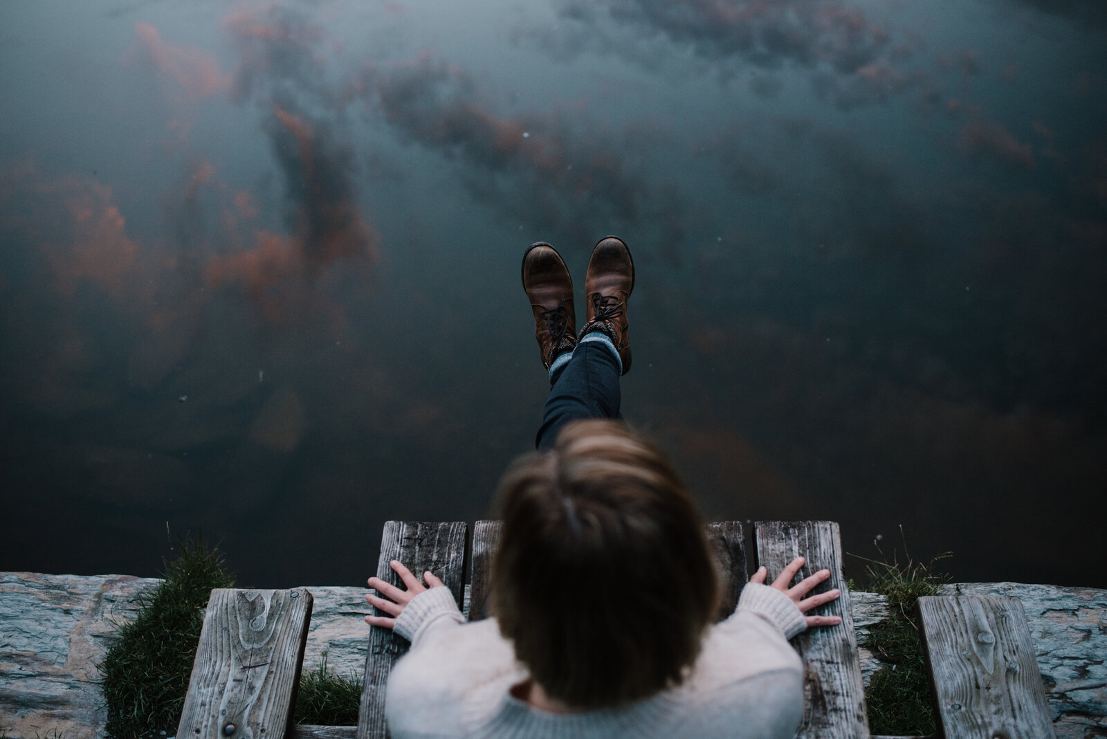 a woman sits on the edge of a pontoon, her feet dangling above the water that is reflecting pink and grey clouds