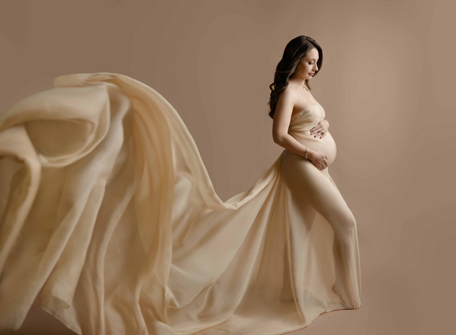 Mom wrapped in cream drape holding baby bump with drape tossed in air. Maternity Portrait by Jenni Nikki Photography at Atlanta maternity photo studio.