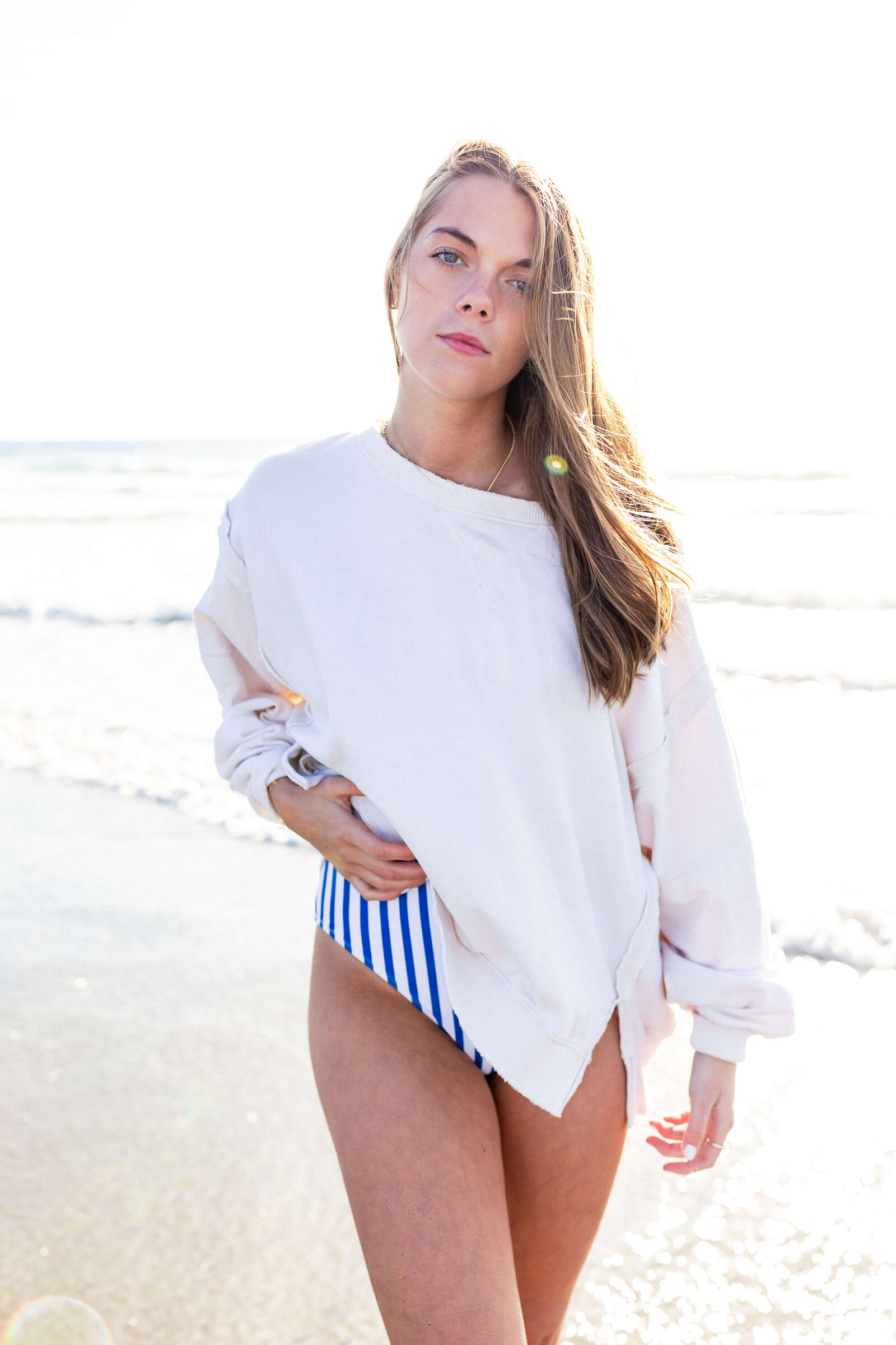 brunette high schooler posing on a beach wearing a white shirt and blue striped swim suit bottoms