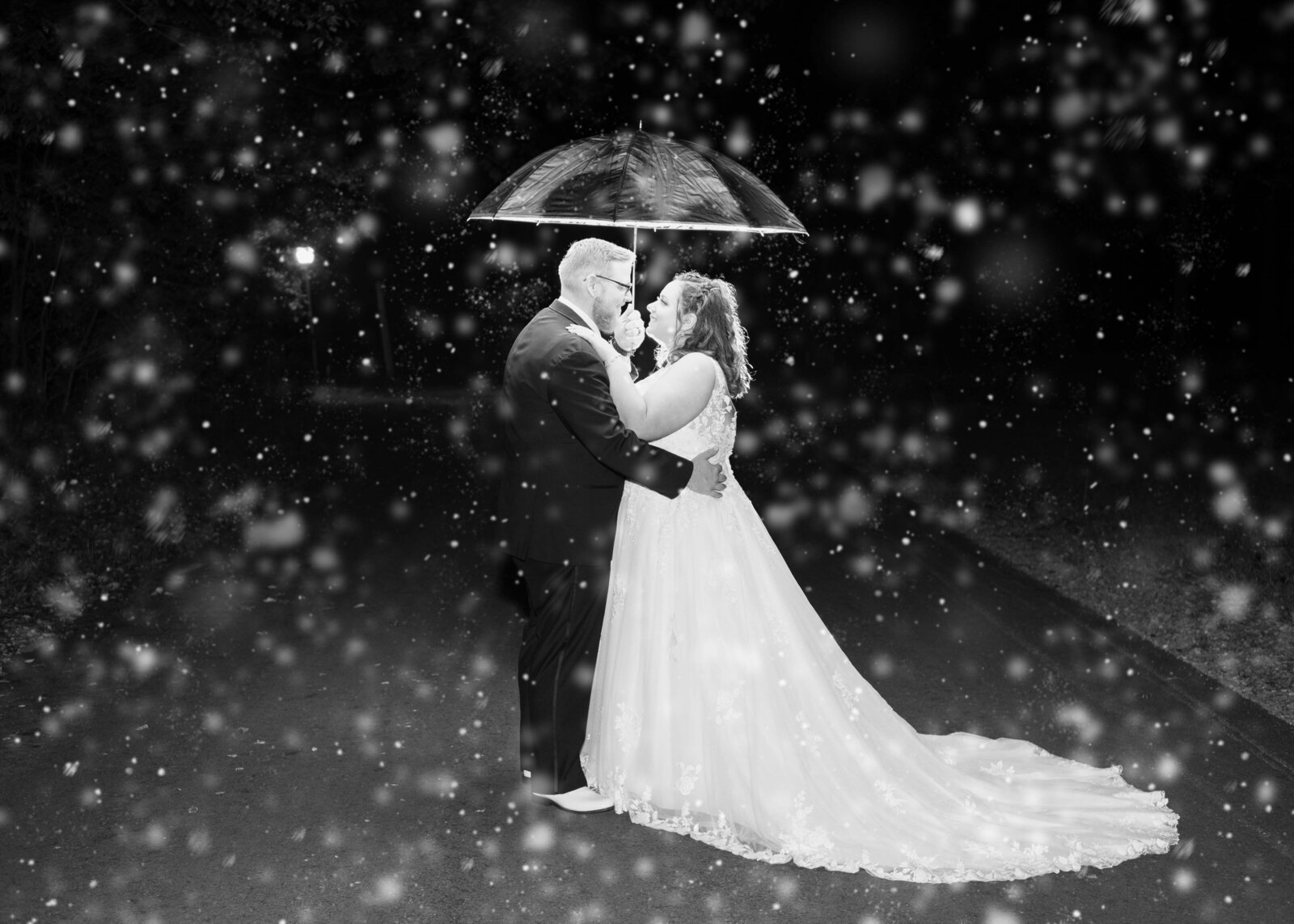 The bride and groom stand under an umbrella at night with a magical falling snow effect, captured during a Blue Knob, PA wedding, blending romance and creative photography.