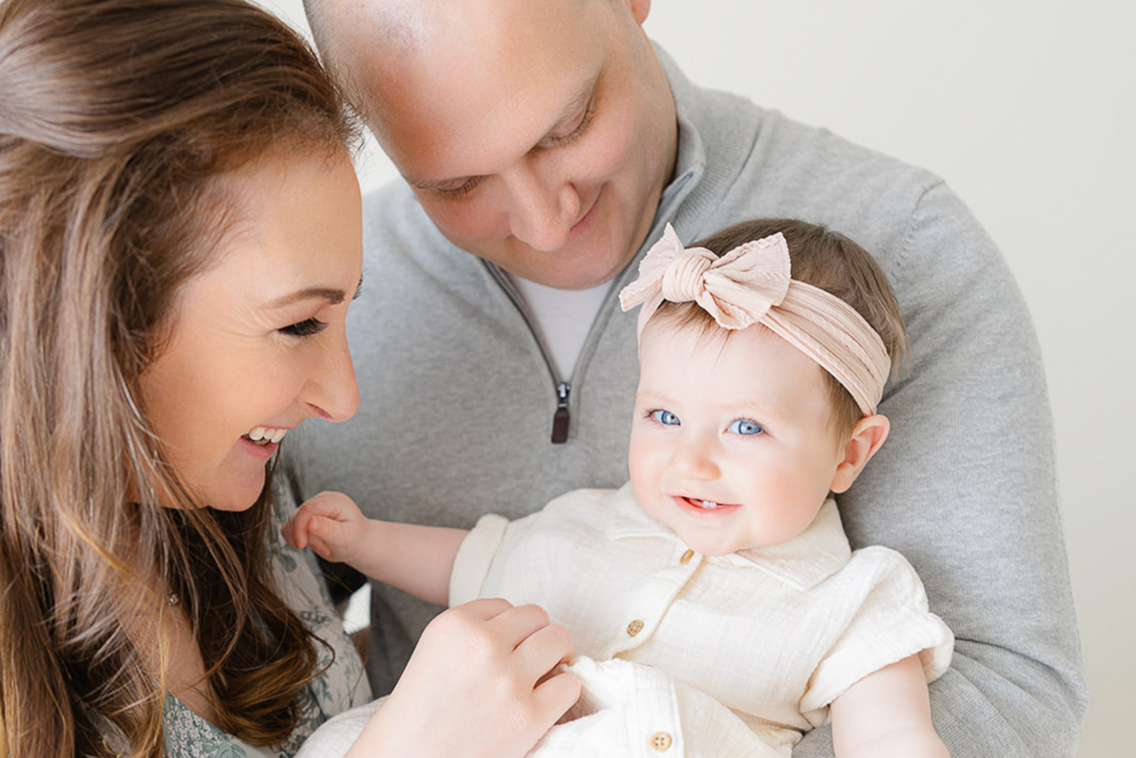 8 month old baby girl wearing pink bow smilies while being held by her parents during boston family photos