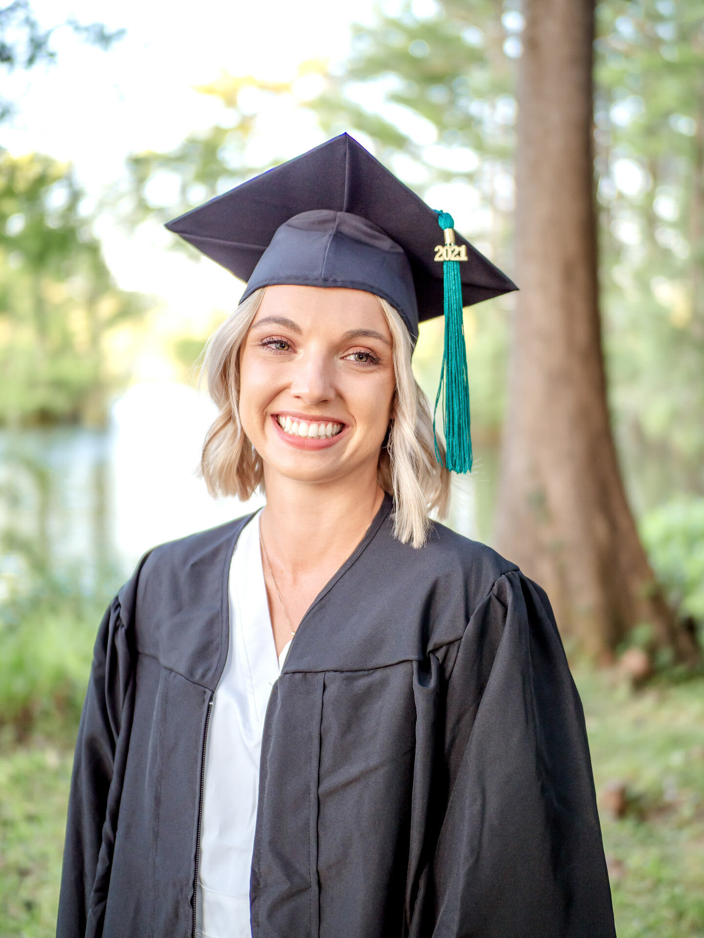 A smiling college senior in a graduation cap and gown standing by a lakeside in Wilmington, North Carolina. This portrait captures the joy of graduation, perfect for those looking for professional senior photography in a natural setting.
