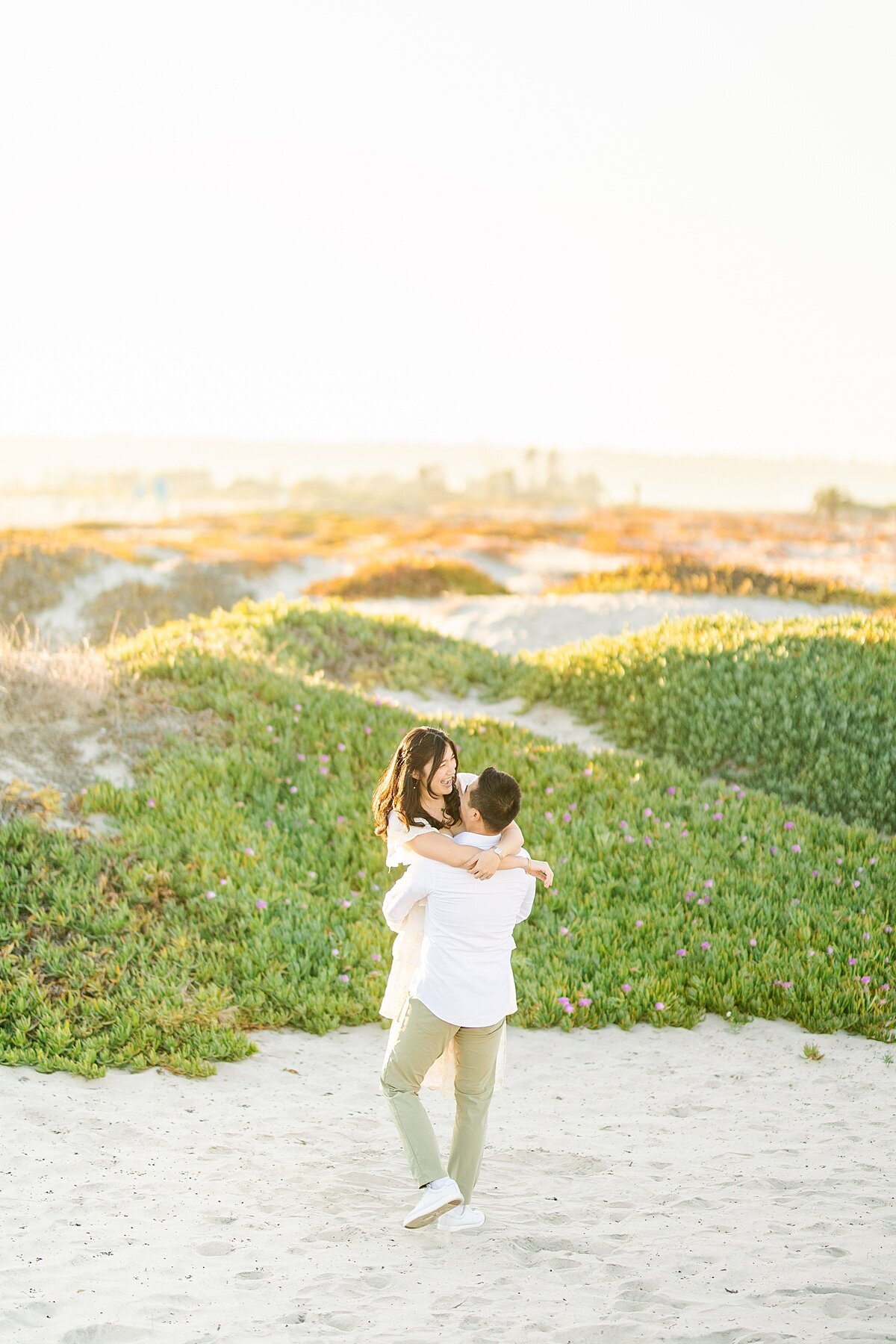 Groom picking up and twirling bride on the san dunes while the sun sets on the beach.