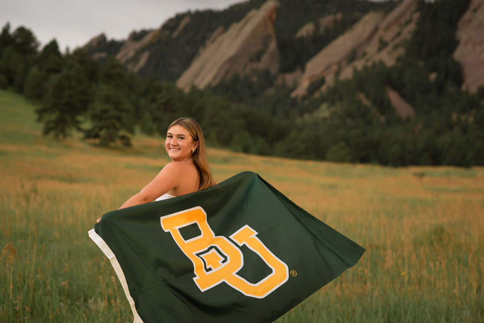 High school senior girl holding a Baylor University flag in a Colorado meadow with the Flatirons in the background during an outdoor senior photography session.