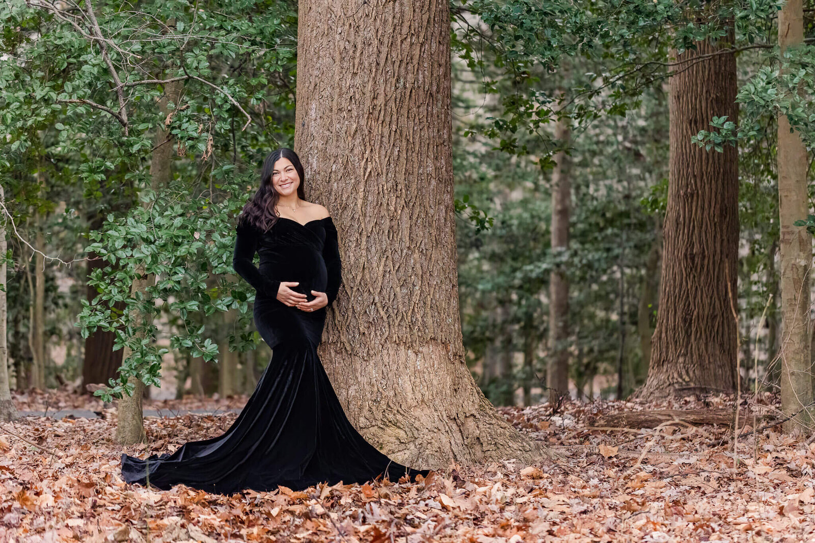 A pregnant woman in a black dress leaning against a tree at a park.