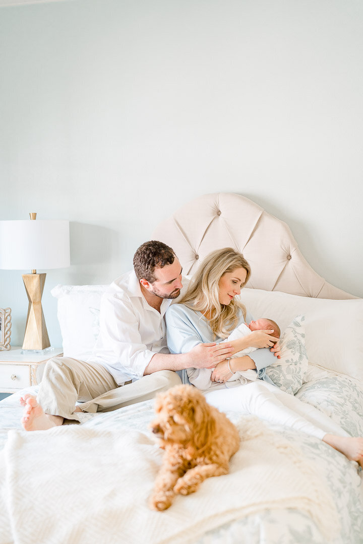 A couple sits on bed holding newborn baby while dog looks on
