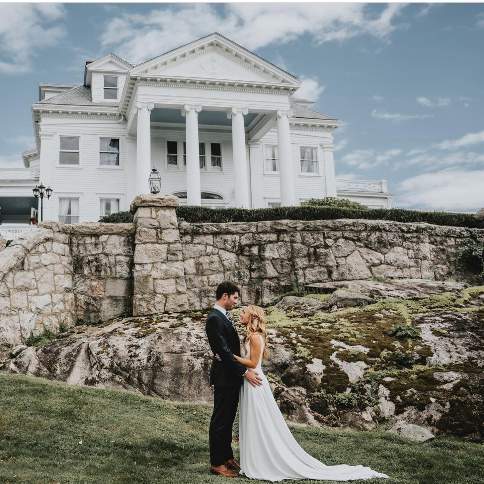 bride and groom standing and holding each other on a grassy area with rocky terrain in front of a large, white columned mansion
