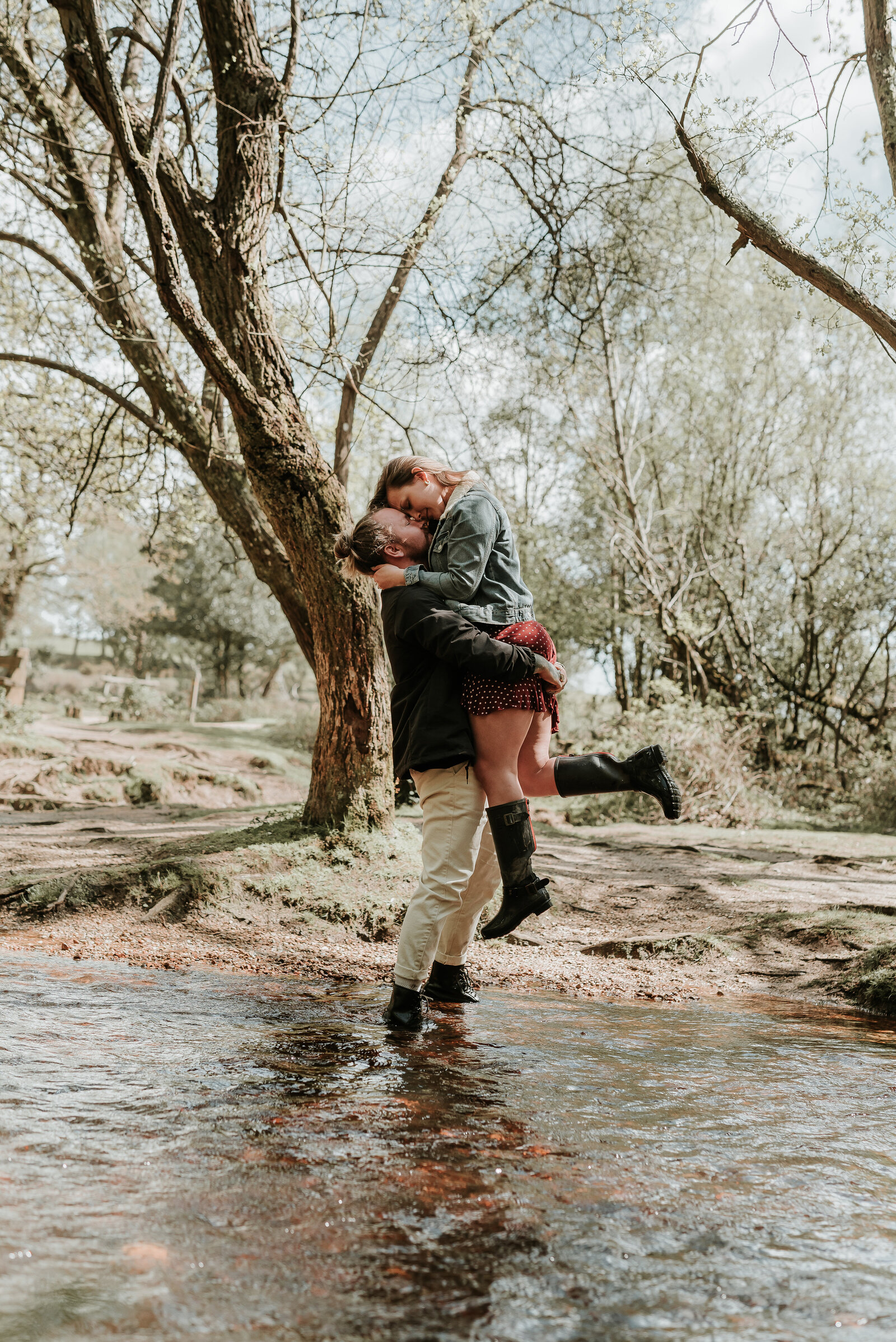 Man lifts his fiance up and couple embrace in a shallow river in the New Forest