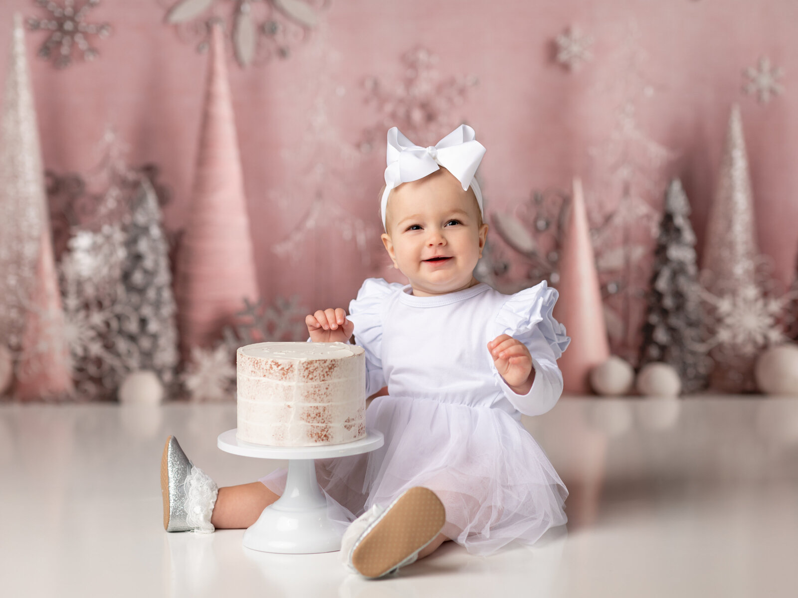 one year old girl in white dress with pink winter theme backdrop for first birthday photos