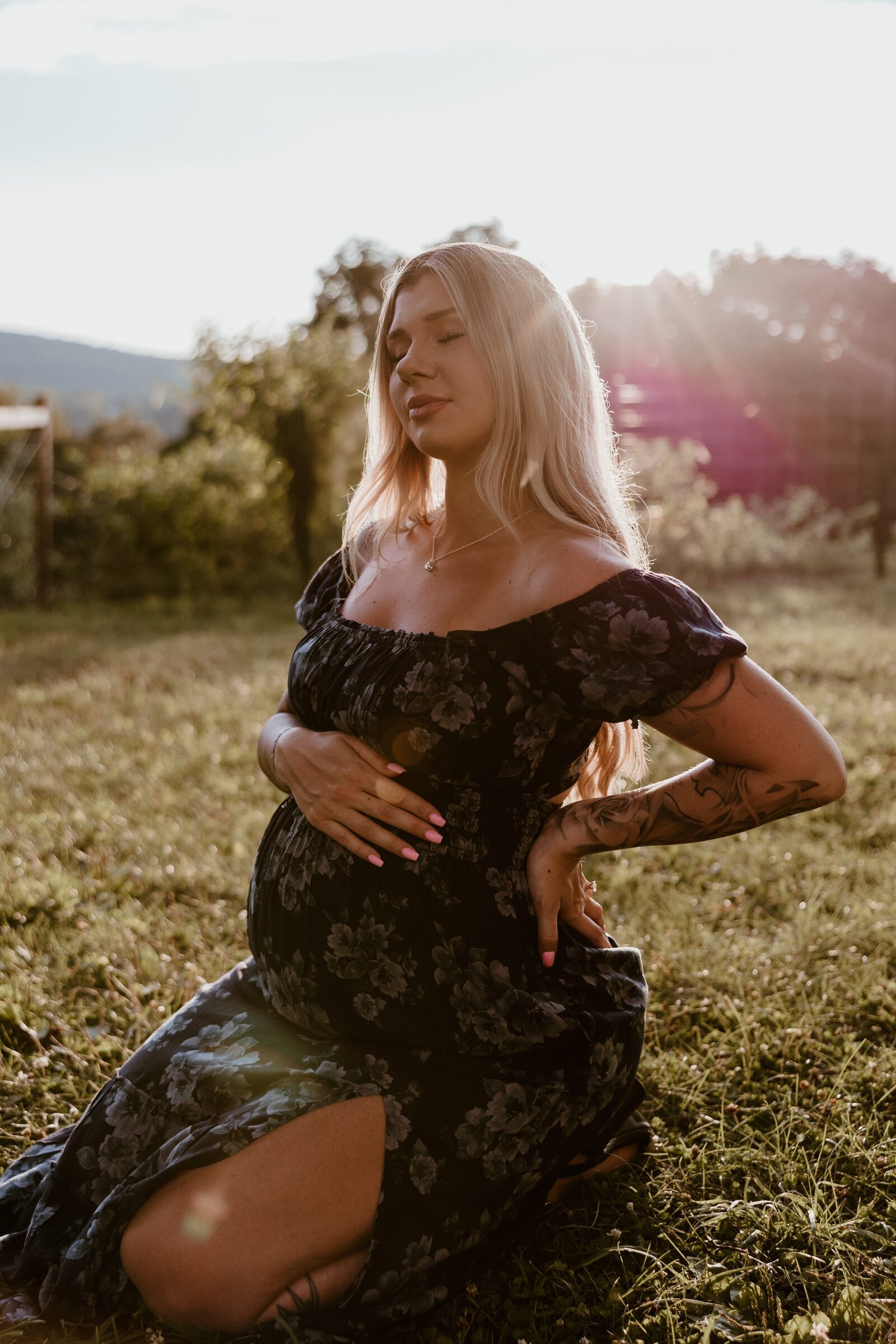 Pregnant woman in a floral dress cradling her belly while sitting in a sunlit field, captured by a Hudson Valley maternity photographer.