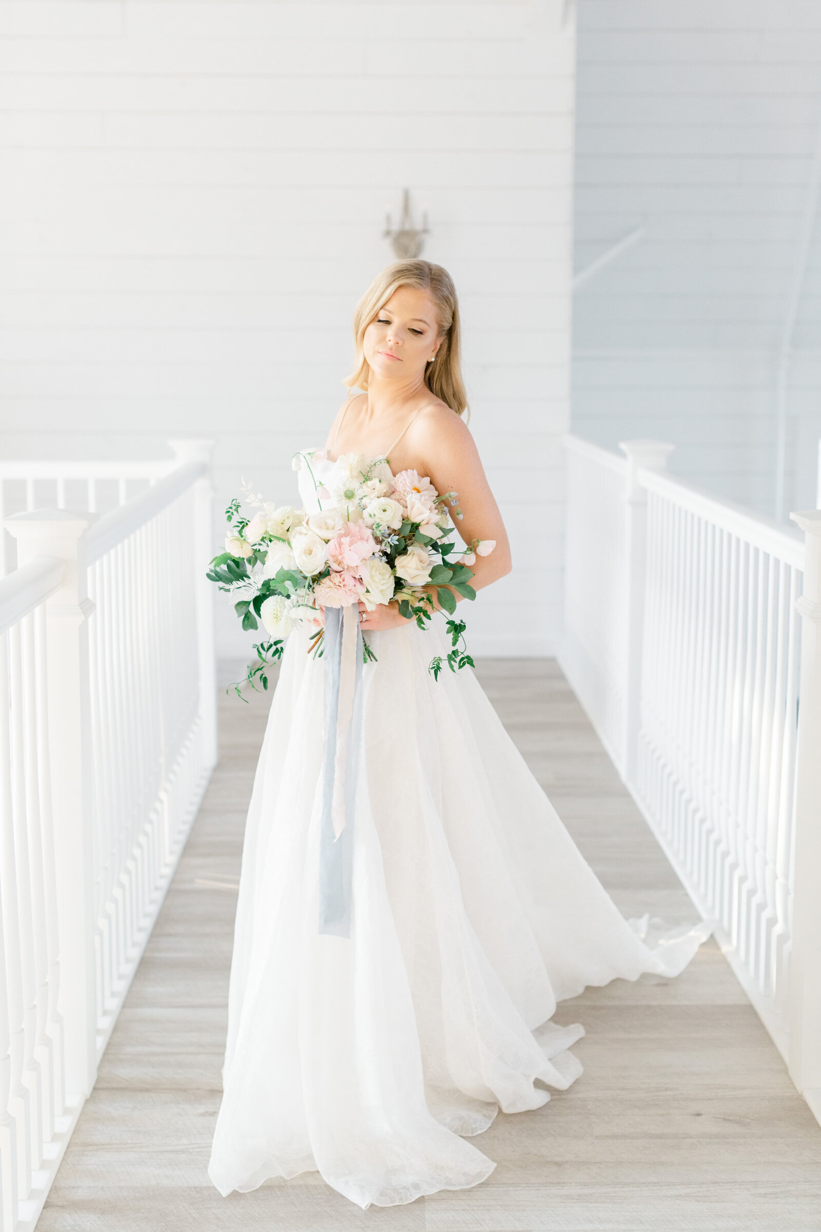 Bride stands on balcony of white barn wedding venue holding her bouquet