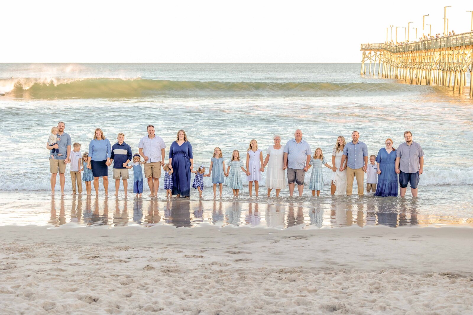 A large family stands together by the ocean near a pier, dressed in coordinating blue and white outfits. The setting sun casts a warm glow, creating a picturesque scene, ideal for professional family photography in NC, showcasing family unity.