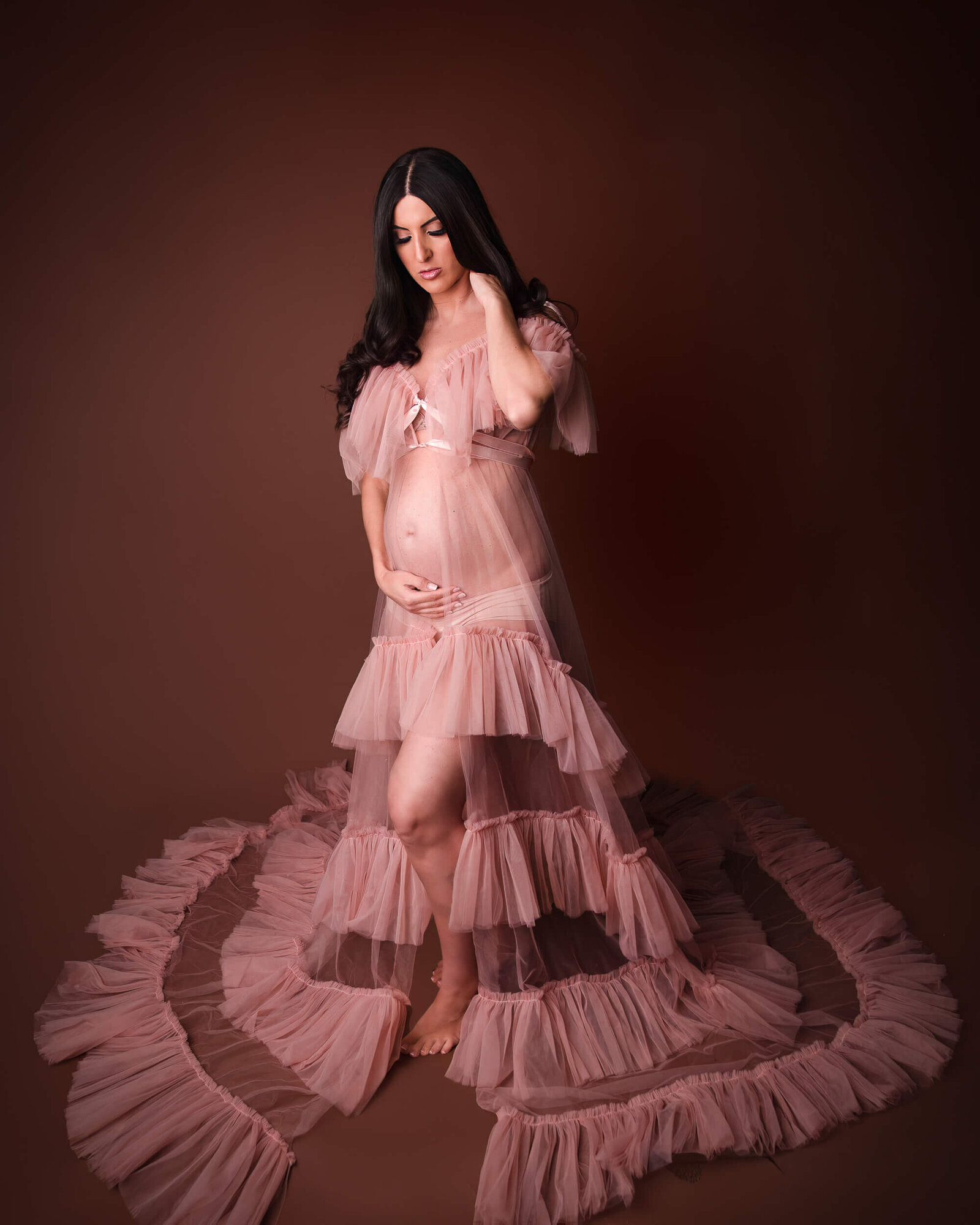 An expectant mother stands elegantly in a ruffled pink tulle gown, the long train fanning out around her, evoking a sense of romance and grace.