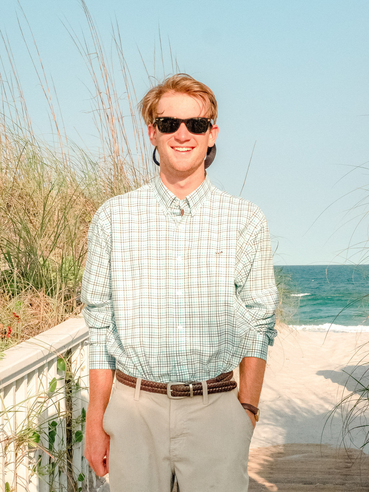 A high school senior wearing sunglasses and a checkered shirt, standing on a Wrightsville Beach path with the ocean in the background in North Carolina. This vibrant image highlights the relaxed and casual vibe of senior photography.
