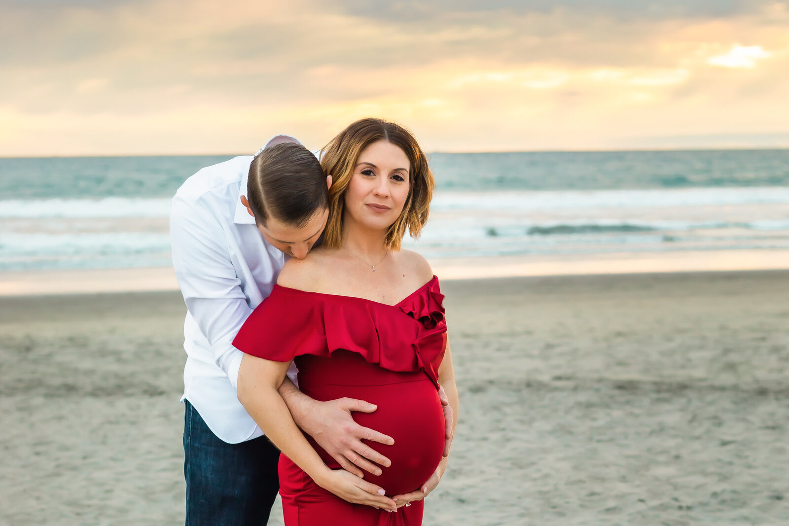 Maternity Photographer, a husband embraces his pregnant wife on the beach at sunset