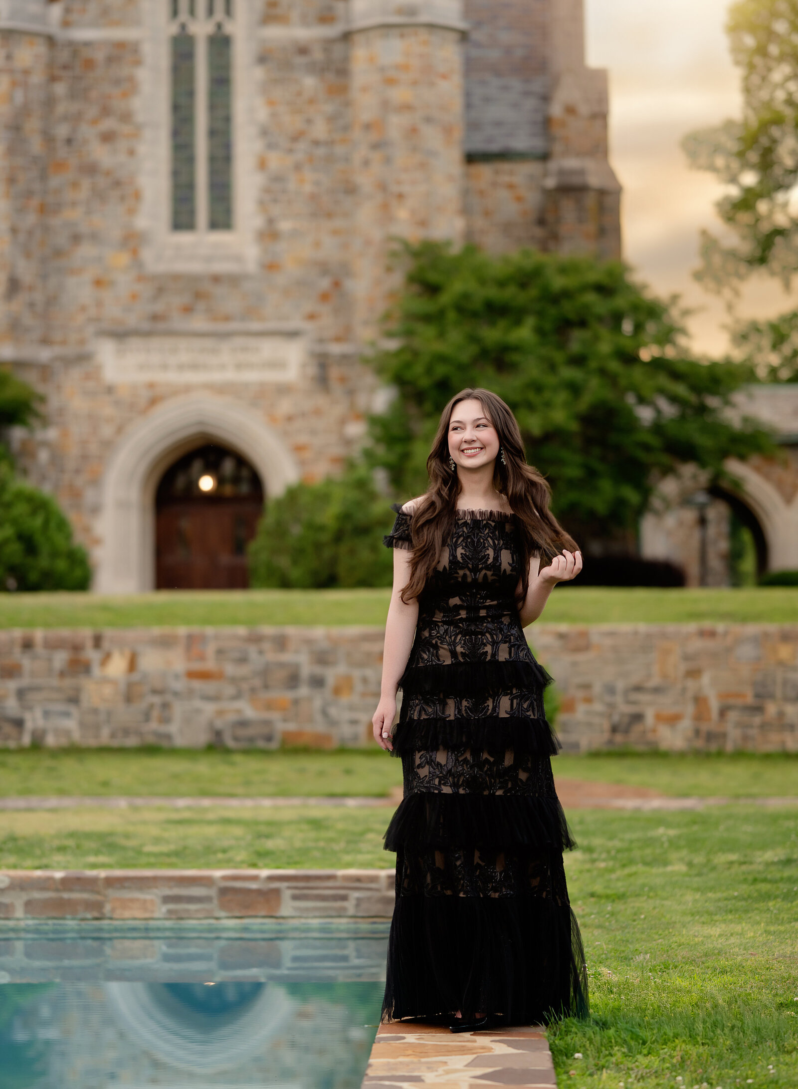 teen girl in black lace dress, playing with hair as she walks next to the reflection pond at Berry College, taking graduation photos.