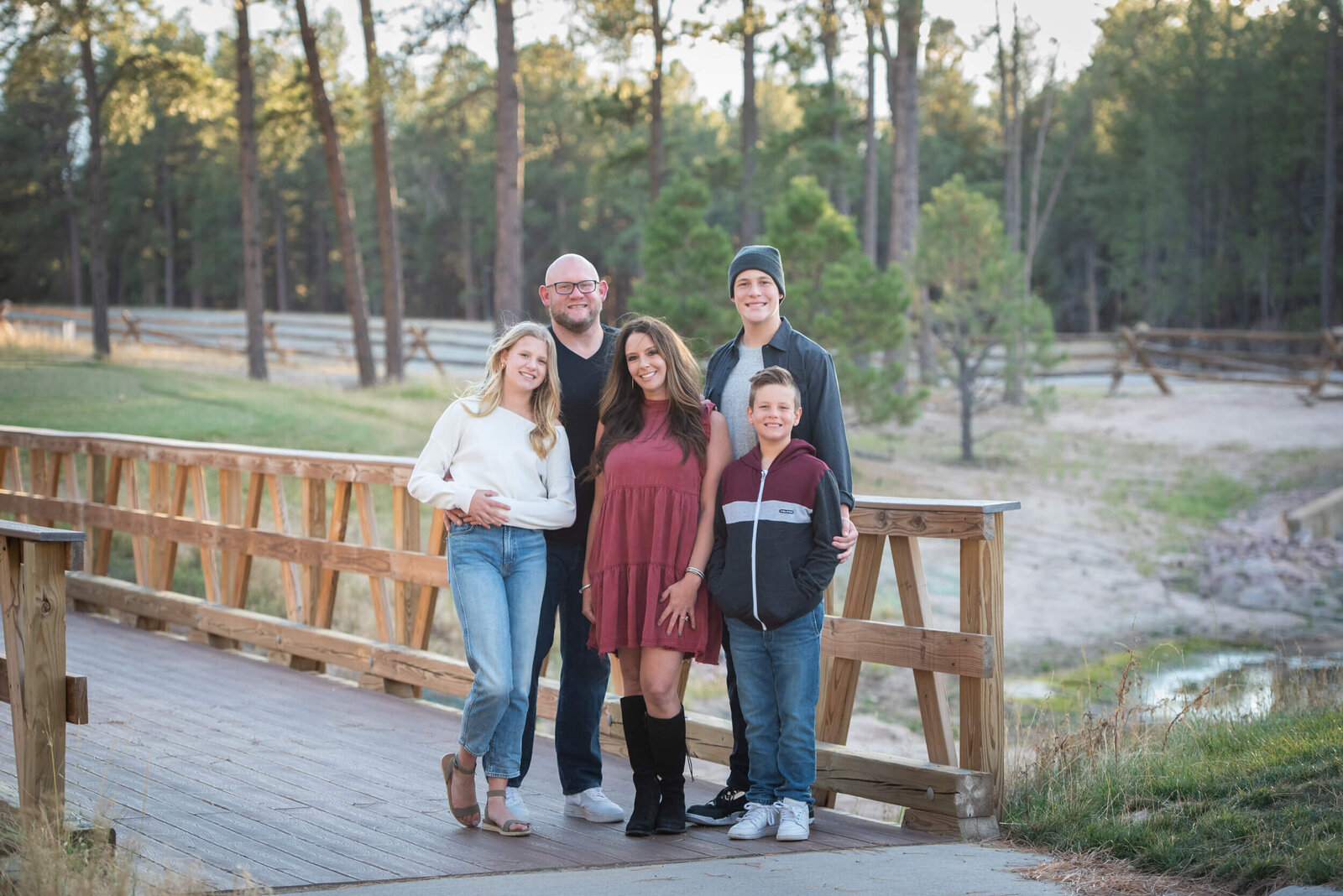 A family of 5 lean on each other and a bridge railing while standing together at sunset in a park