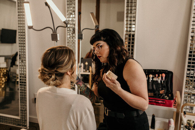 a behind the scenes look of a bride getting her makeup applied before her wedding.  Bride is in a robe sitting in a chair while the dark haired makeup artist stands in front of her applying her makeup.