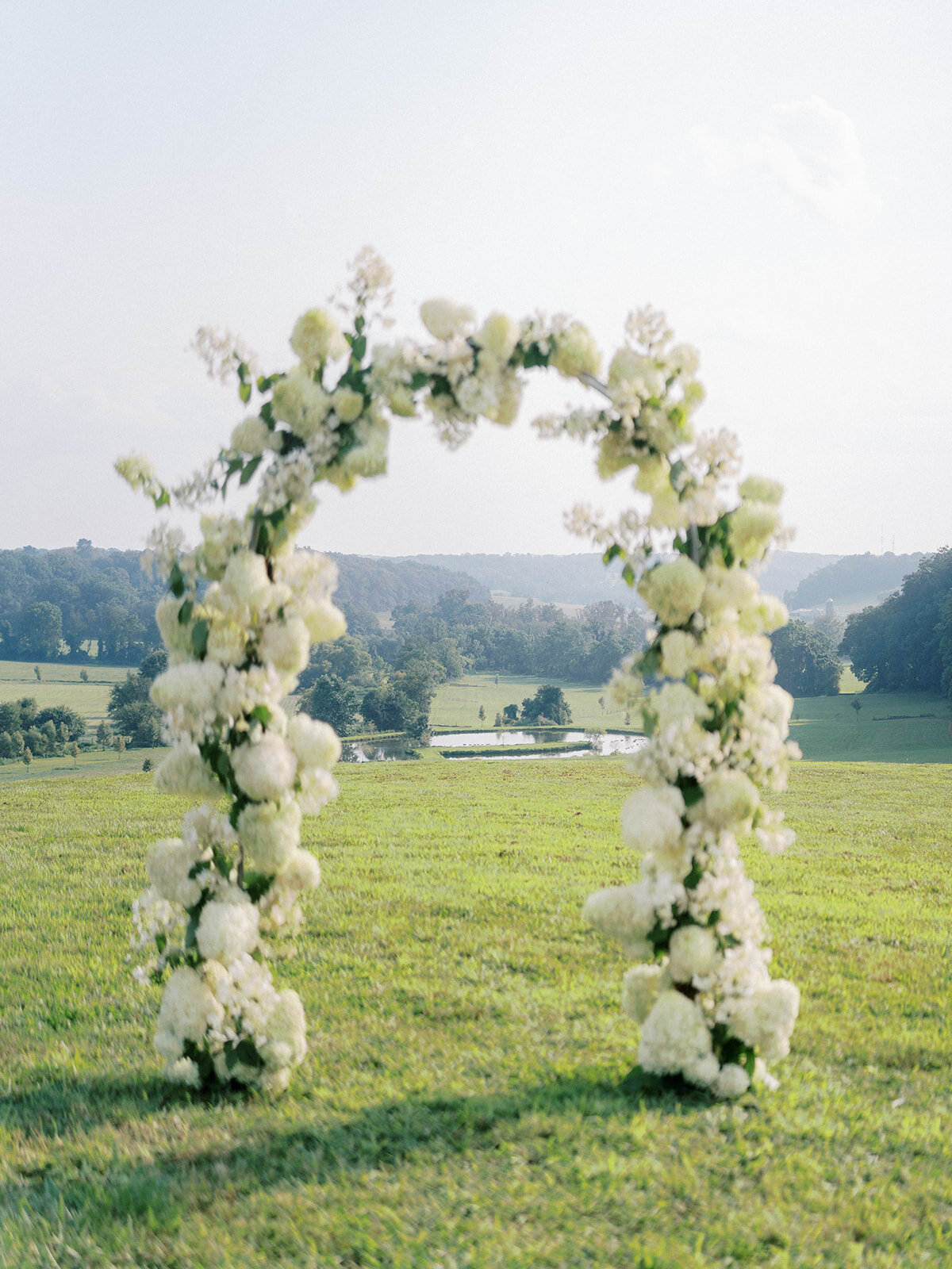 Full coverage floral arch filled with white airy quick-fire hydrangea and limelight hydrangeas overlooking the landscape at Harford Hills wedding venue in Fallston Maryland rolling hills.