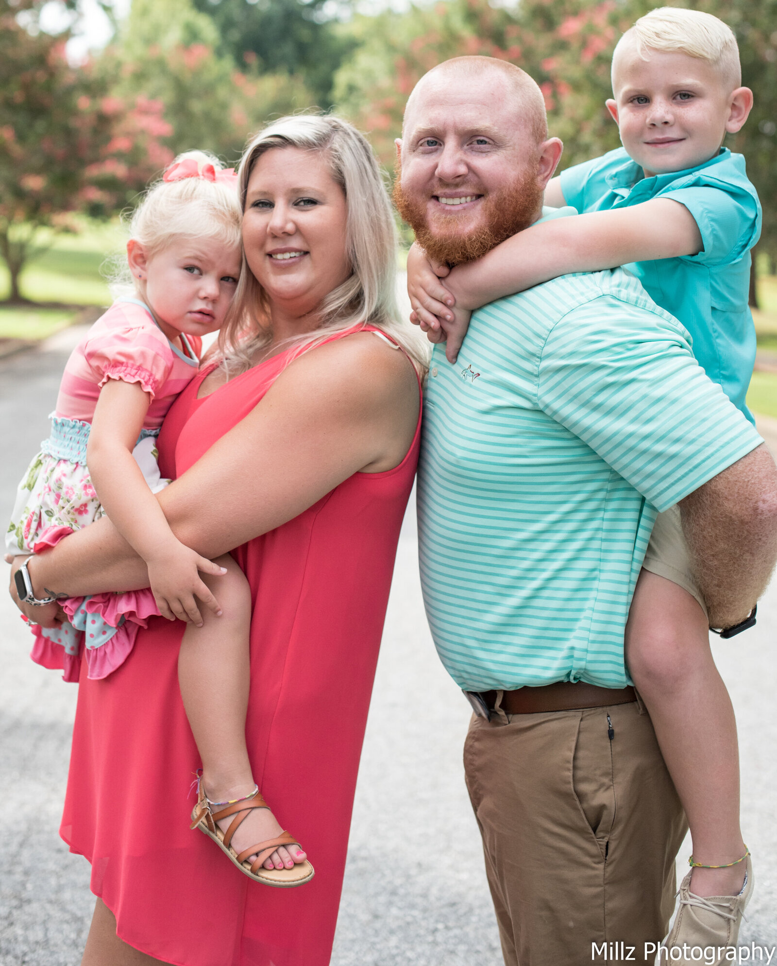 a family of four, mother holding her daughter and father carrying his son on his back, standing outside smiling at the camera photographed by Millz Photography in Greenville, SC