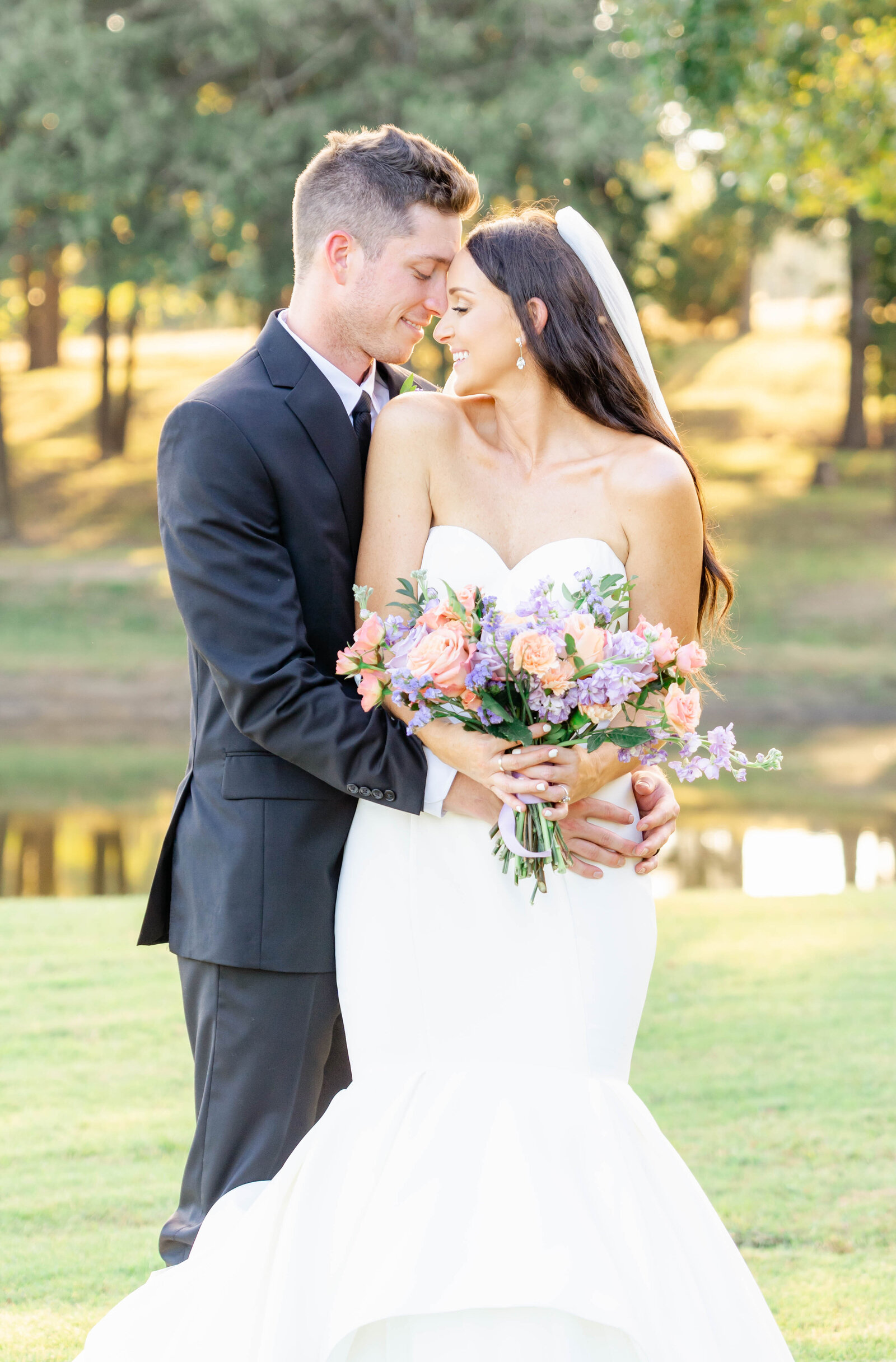 Portrait of bride and groom in a white wedding gown and black tuxedo embracing each other in front of a body of water.