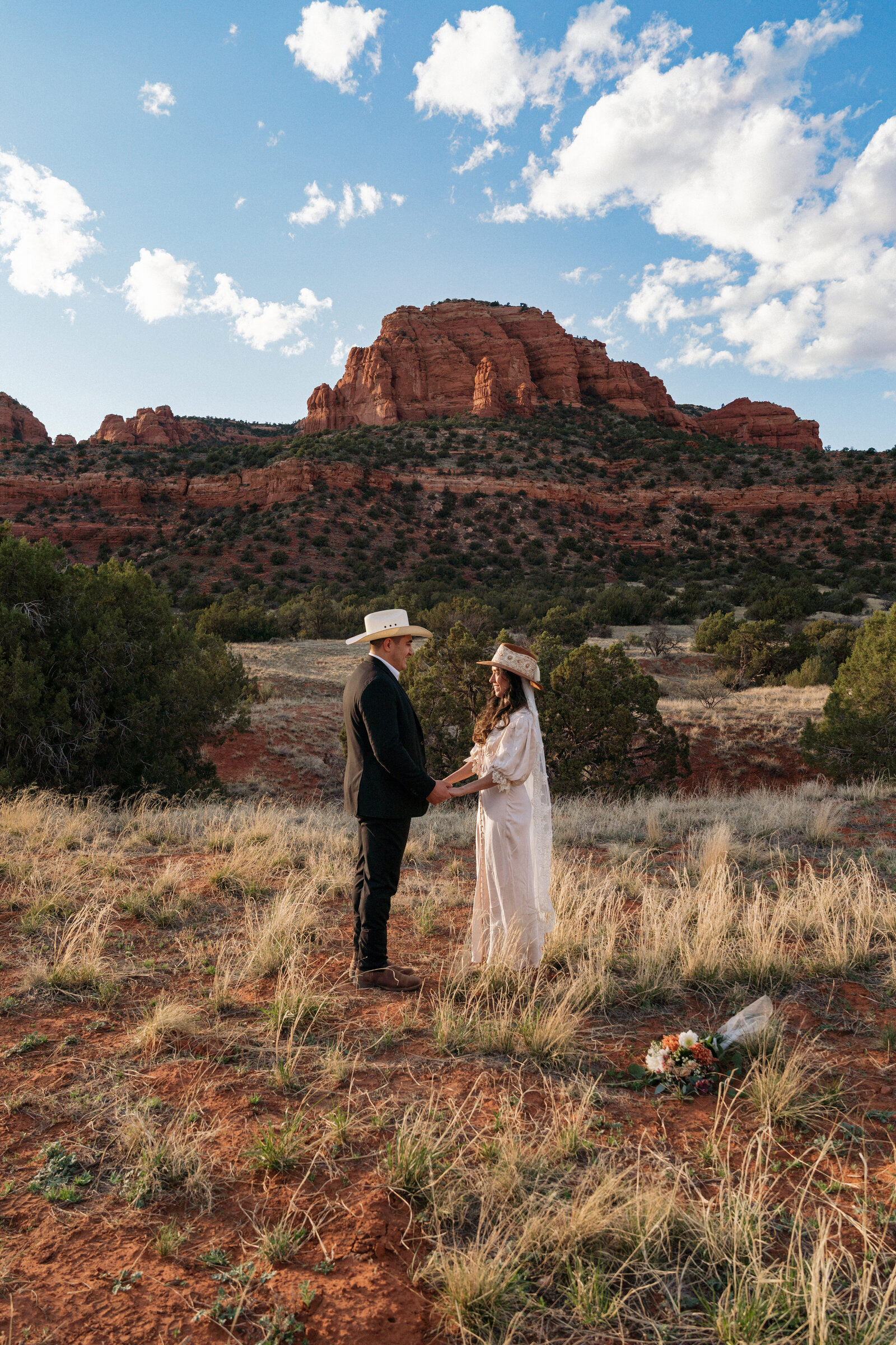 Bride and groom holding hands in western attire during their Sedona elopement ceremony at Bear Mountain, surrounded by red rocks and desert landscape.