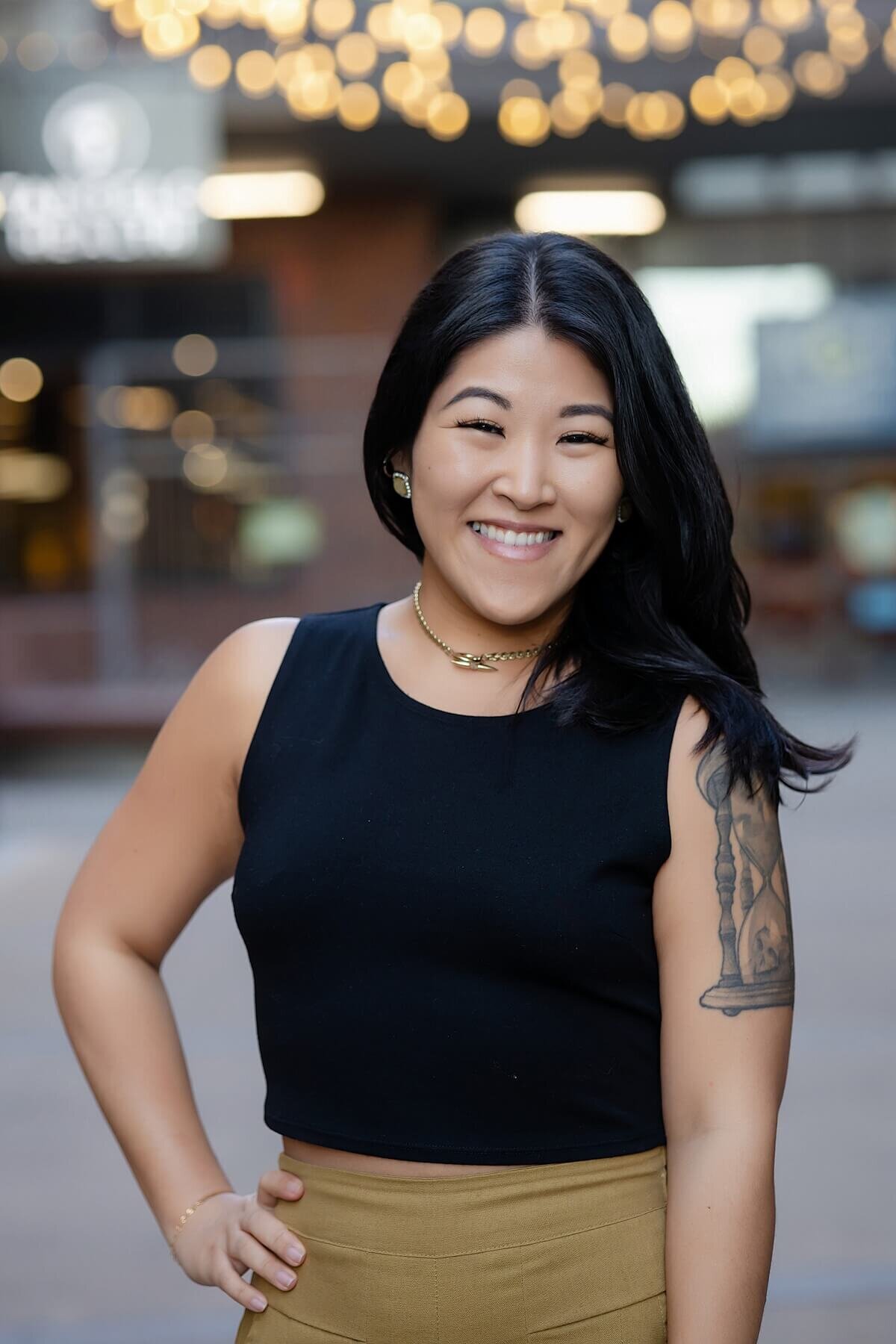 Professional branding photo of a smiling Asian woman in a black sleeveless dress, standing confidently in an urban Seattle setting. Featuring bold tattoos and a polished look, this image captures personal brand power and authenticity during a Seattle branding photoshoot for women in business.